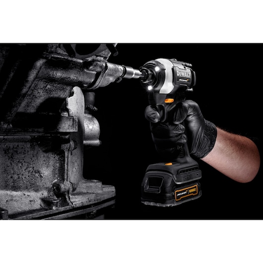A person wearing a black glove uses a DEWALT cordless impact driver to tighten or loosen a bolt on a metallic machine part. The tool is equipped with LED lights illuminating the work area.