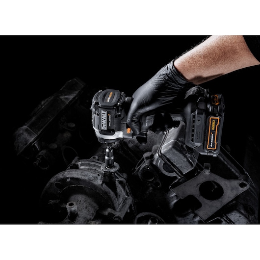 A person wearing a black glove uses a DEWALT cordless impact wrench to work on a metal machinery part against a dark background.