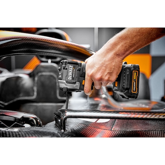 A person uses a DEWALT cordless impact driver (model DCF85ME2GT) to fasten a screw into a carbon fiber automotive part, likely part of a racing vehicle.