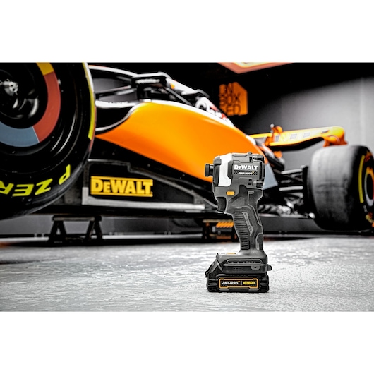 A DEWALT cordless impact driver (model DCF85ME2GT) is placed on a concrete floor in front of an orange and black race car in a garage setting.