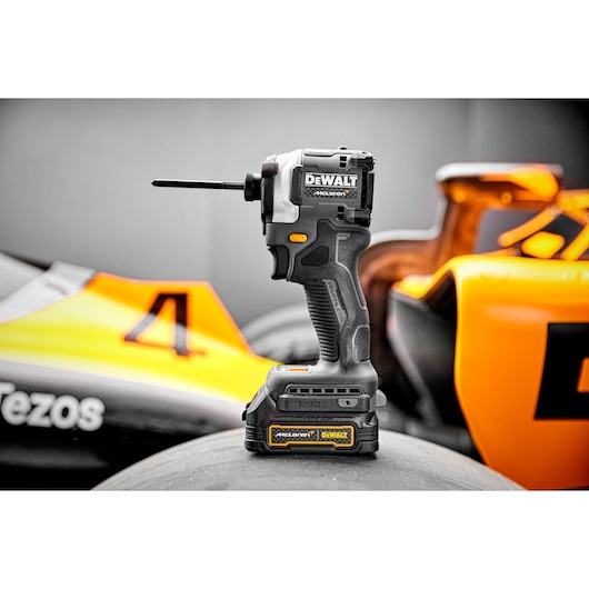 A DEWALT cordless impact driver tool is shown standing upright on a tire, with a racing car featuring orange and yellow details in the background.