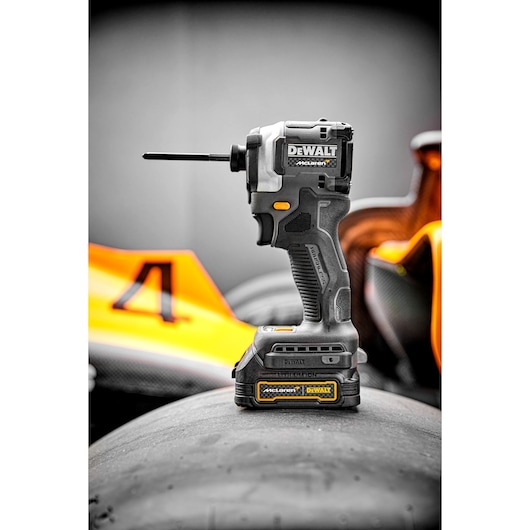 A DEWALT cordless impact driver, model DCF85ME2GT, is standing upright on a racing car tire, with a racing car featuring orange details and the number 4 in the background.