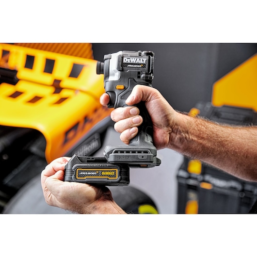 A person is holding a DEWALT McLaren cordless power tool, attaching a rechargeable battery pack to the bottom of the tool. The background shows yellow and black DEWALT branded equipment.