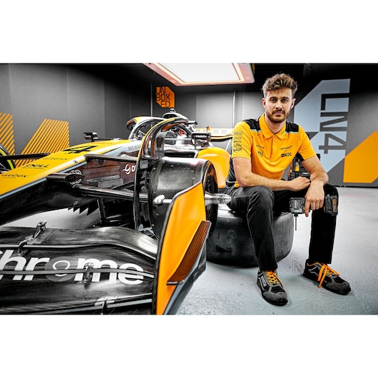Person in orange shirt with DEWALT tool sits by a yellow race car in a modern garage.