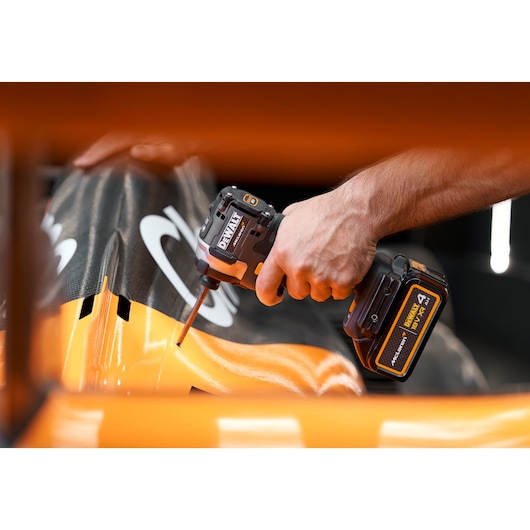 A DEWALT cordless impact driver is used on an orange and black surface, close-up of hand and tool in action.