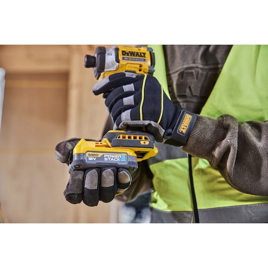 Worker holding a DEWALT 18V Powerstack cordless tool, wearing gloves and safety vest at construction site.
