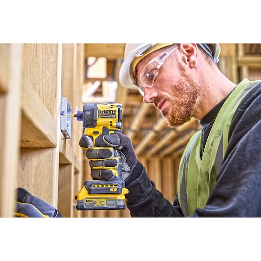 Worker uses DEWALT DCF870E2T impact driver on a wood frame, wearing gloves, helmet, and safety vest.