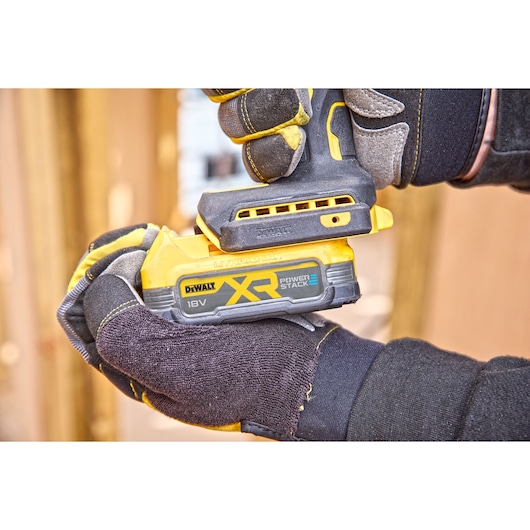 Gloved hands hold a DEWALT XR POWERSTACK 18V cordless tool, showing its battery and grip in a workshop setting.
