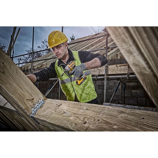 Person using a DEWALT cordless drill on wooden beams at a construction site, wearing safety gear and high-visibility vest.