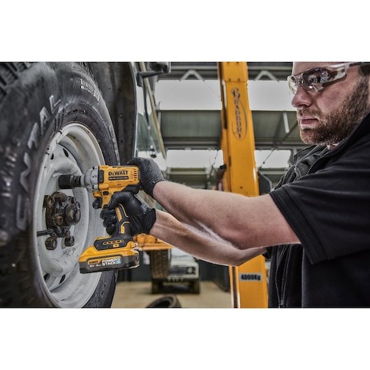 A person wearing black gloves uses a DEWALT DCF891H2T cordless impact wrench to tighten or loosen the lug nuts on a vehicle wheel in a garage setting.