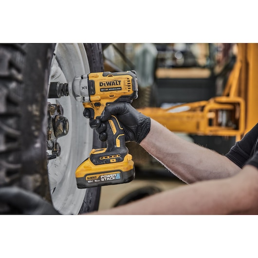 A person using a DEWALT DCF891H2T cordless impact wrench to loosen bolts on a large vehicle wheel. The tool is yellow and black, and the user is wearing black gloves.