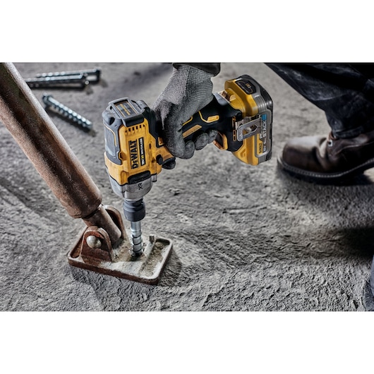 A person wearing work gloves and boots using a DEWALT cordless impact wrench to tighten a bolt on a rusty metal bracket attached to a concrete surface.