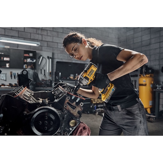 A person with their face blurred is using a DEWALT DCF891H2T cordless impact wrench to work on an engine block in a workshop setting. The workspace includes tools and equipment, with a DEWALT air compressor visible in the background.