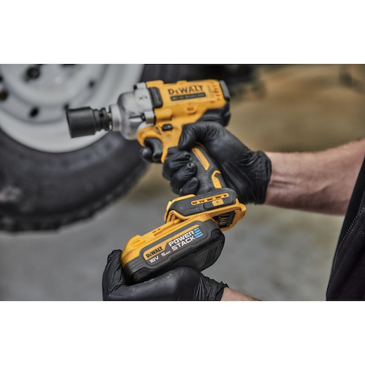 Close-up image of a person wearing black gloves holding a DEWALT cordless impact wrench with a POWERSTACK 18V 5.0Ah battery attached, in front of a vehicle wheel.