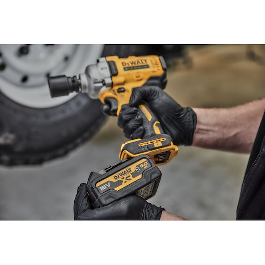 Gloved hands hold a DEWALT cordless impact wrench and an 18V battery in front of a blurred tire.