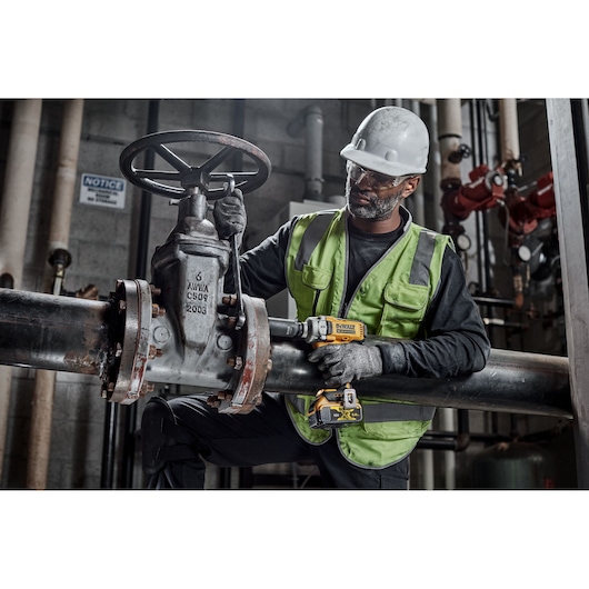 Worker uses DEWALT DCF892P2T impact wrench on industrial pipes, wearing safety gear and gloves.
