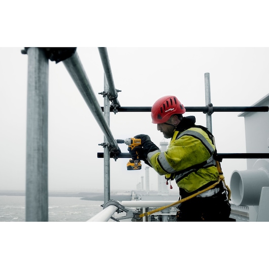 Worker with safety gear uses DEWALT tool on metal pipes outdoors in industrial setting.