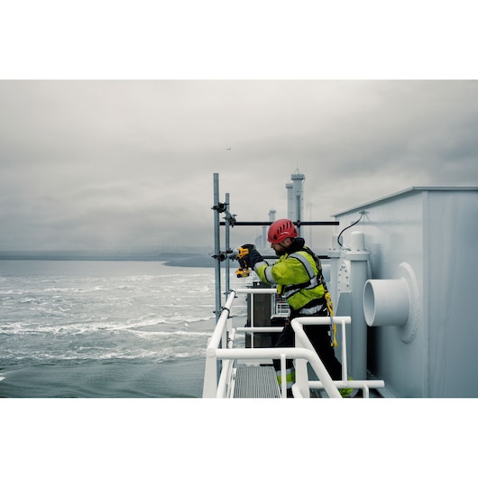 Worker in safety gear uses DEWALT tool outdoors near water and metal railings on an industrial platform.