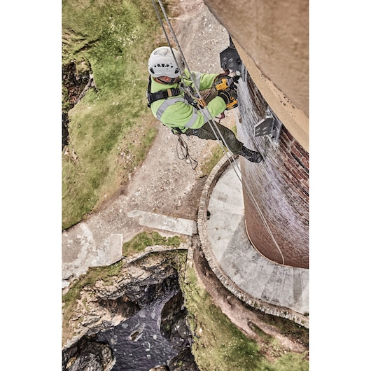 Worker with safety gear using a DEWALT tool while rappelling on the side of a tall brick structure outdoors.