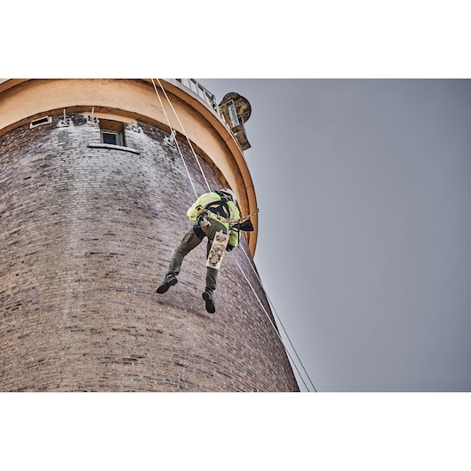 Worker rappelling down a tall brick tower with safety harness, holding DEWALT tool.