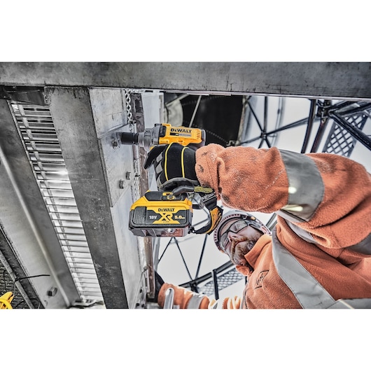 Worker uses a DEWALT cordless impact wrench on metal structure outdoors, wearing safety gear and gloves.