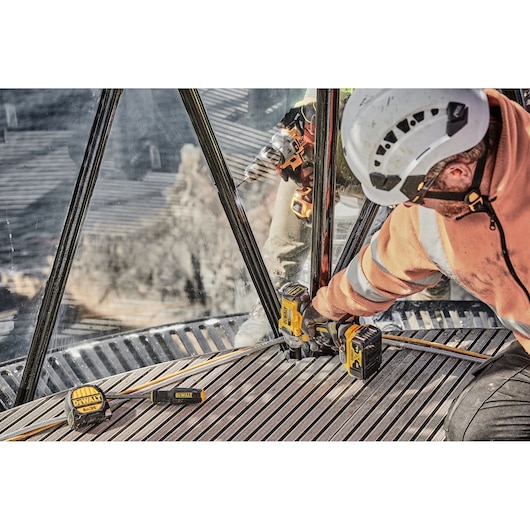 Worker uses DEWALT DCF894P2 impact wrench on metal structure. DEWALT tools and battery are visible on site.