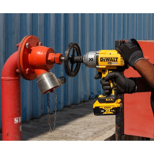 Person uses a DEWALT cordless impact wrench to turn a valve on a red outdoor hydrant beside a blue wall.