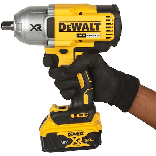 A person wearing a black glove holds a DEWALT DCF899P2-QW 18V XR brushless cordless impact wrench with a 5.0Ah Li-ion battery, shown against a white background.
