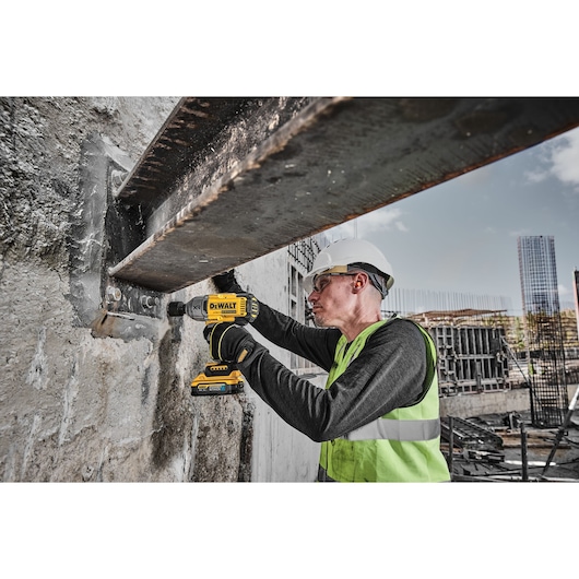 Worker using a DEWALT DCF900H2T drill on a steel beam at a construction site, wearing safety gear and helmet.