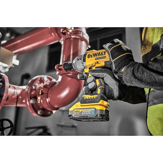 A gloved hand uses a yellow DEWALT cordless impact wrench near large red industrial pipes indoors.