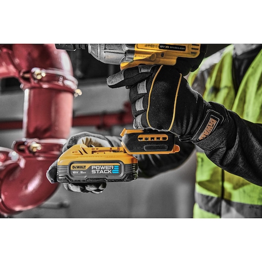 Gloved hand using DEWALT DCF900H2T power tool with POWERSTACK battery near industrial pipes.
