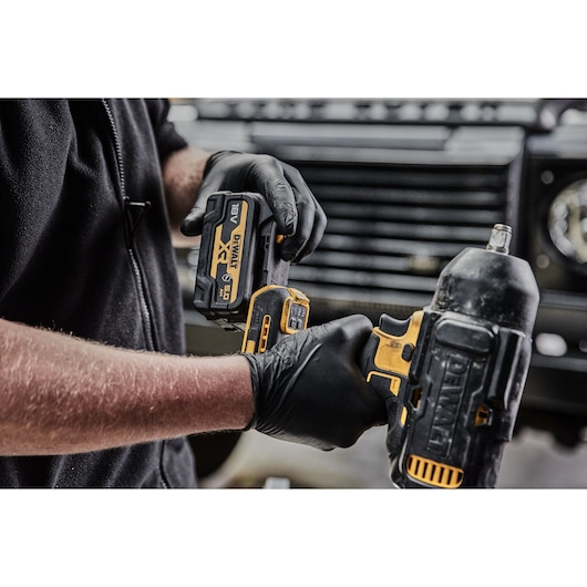 Gloved hands attaching a DEWALT battery to a cordless impact wrench, with a vehicle grille in the background.