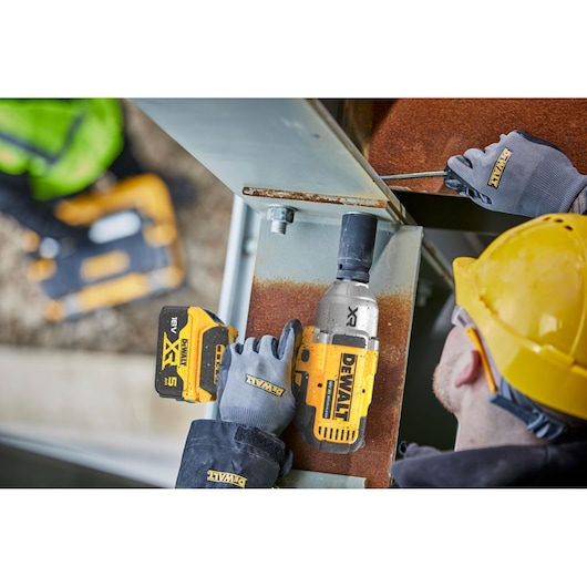 Worker uses DEWALT cordless impact wrench on metal beam while wearing gloves and yellow helmet.