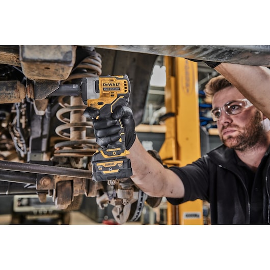 A person using a DEWALT cordless impact wrench (DCF901P1G) to work on the underside of a vehicle in a workshop. The person is wearing black gloves and the tool is being applied to a bolt.