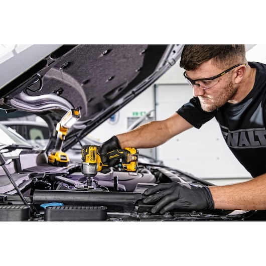 A person wearing black gloves uses a yellow DEWALT cordless impact wrench (SKU: DCF902D2) to work on the engine of a car. An additional DEWALT work light is positioned under the raised hood for illumination.