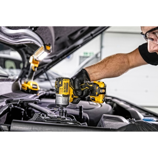 A person wearing gloves uses a DEWALT DCF902D2 cordless impact wrench to work on a car engine, with a DEWALT work light illuminating the area.