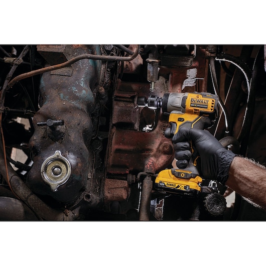A person wearing a black glove uses a DEWALT DCF902D2 cordless impact wrench to work on a part of an engine or machinery. The tool is yellow and black and is being applied to a rusty metal component.