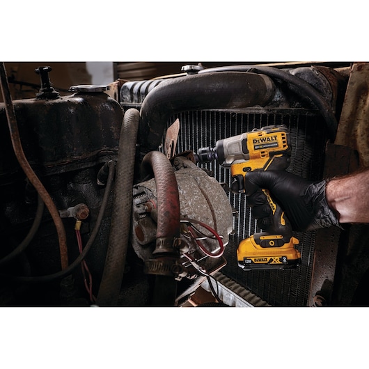 A person using a DEWALT DCF902D2 cordless impact wrench to work on a part of a vehicle engine. The person is wearing a black glove and the tool is yellow and black.