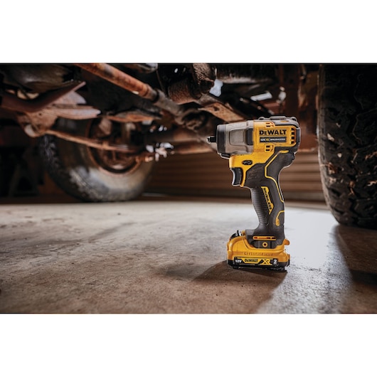 A DEWALT DCF902D2 cordless impact wrench with a yellow and black design is shown standing upright on a garage floor, positioned beneath a vehicle with visible suspension and tire components.