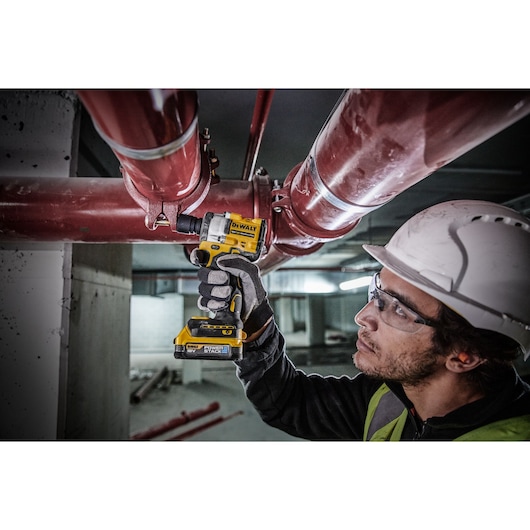 A person wearing a safety helmet and gloves uses a DEWALT cordless impact wrench (model DCF921E2T) to tighten a bolt on a large red pipe in an industrial setting. The person's face is blurred for privacy.