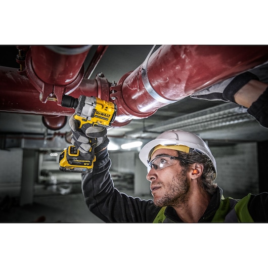 A person wearing safety gear uses a DEWALT cordless impact wrench (model DCF921E2T) to work on large red industrial pipes in an indoor setting. The person's face is blurred.