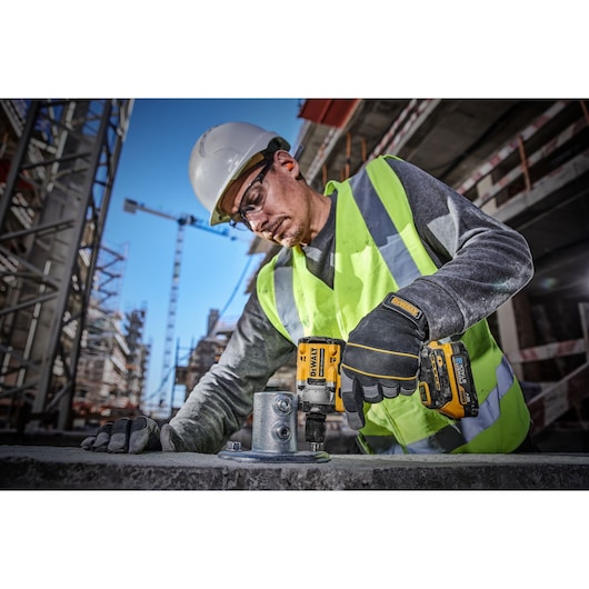 A construction worker wearing a safety helmet, high-visibility vest, and gloves is using a DEWALT DCF921E2T cordless impact wrench to secure bolts on a metal structure at a busy construction site.