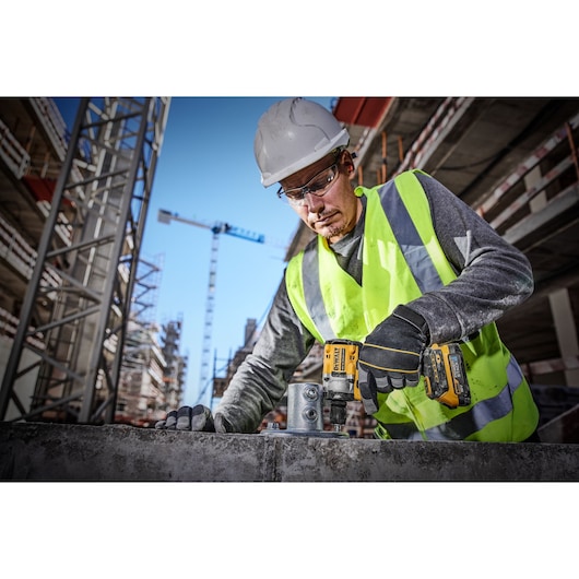 A construction worker wearing a high-visibility vest and safety helmet uses a DEWALT DCF921E2T power tool on a building site. Several cranes and unfinished buildings are visible in the background.