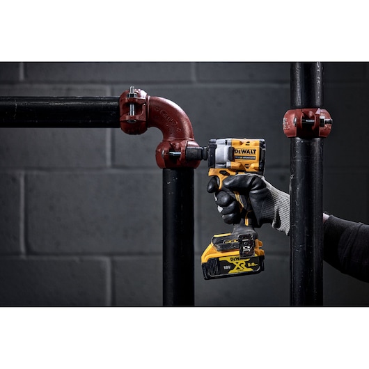 A person wearing black gloves is using a yellow DEWALT cordless power tool to tighten a metal pipe fitting.