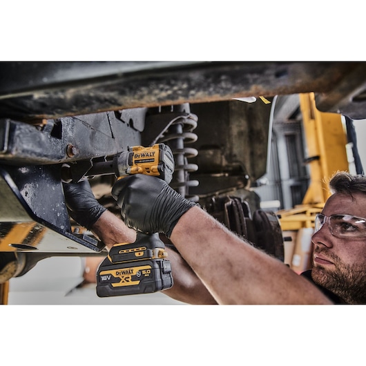A person wearing black gloves uses a DEWALT 18V XR cordless impact wrench to work on a vehicle's suspension system in a workshop.