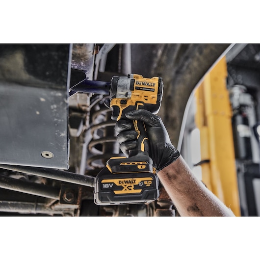 A person wearing a black glove uses a DEWALT cordless impact wrench with a 18V 5.0Ah battery to work on a metal structure, possibly under a vehicle.