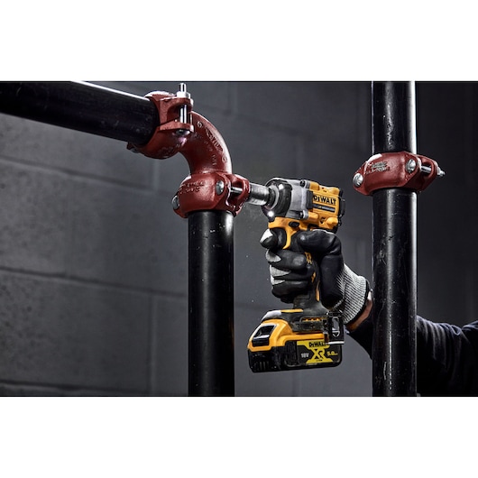 A person wearing work gloves uses a yellow DEWALT cordless impact wrench (model DCF922NT) to tighten a metal pipe fitting in an industrial setting.