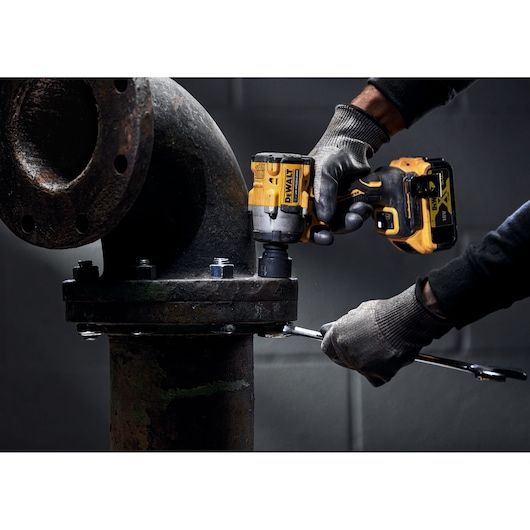 A person wearing gloves is using a DEWALT cordless impact wrench to tighten bolts on a large metal industrial pipe fitting.