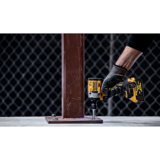 A person wearing a black glove uses a DEWALT cordless impact wrench to tighten a bolt on the base of a metal column, with a chain-link fence in the background.