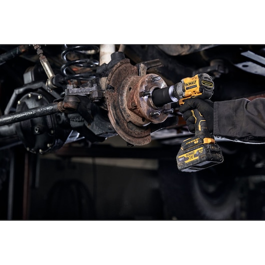 A DEWALT DCF923P2G cordless impact wrench is being used to work on a rusty wheel hub assembly under a vehicle. The tool is held by a gloved hand.